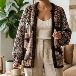 May include: A chunky knit cardigan featuring a brown, beige, and cream geometric pattern with a fuzzy textured trim along the open front. The garment has long sleeves with ribbed cuffs and a relaxed fit, worn over a white ribbed tank top and beige trousers.