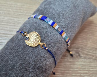 May include: Two bracelets on a gray surface. One bracelet features a hammered gold-tone disc with a blue stone and a dark blue cord. The other bracelet has a beaded design with blue, white, and orange rectangular beads.