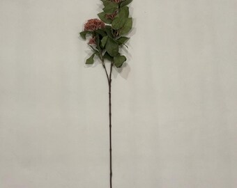 Könnte beinhalten: Ein einzelner künstlicher Blumenstiel mit zwei Büscheln kleiner, rosaroter Blüten und mehreren grünen Blättern. Der Stiel ist dunkelbraun und erstreckt sich nach unten. Der Hintergrund ist eine schlichte, cremefarbene Oberfläche.