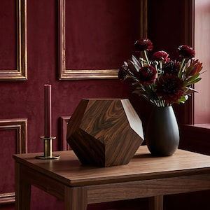 May include: A wooden geometric vase with a dark finish sits on a wooden table, next to a black vase holding dark red flowers. A burgundy candle in a gold holder is also on the table. The background is a deep red wall.
