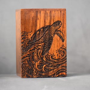 May include: A wooden box featuring a laser-etched design of a sea turtle above ocean waves. The box is made of reddish-brown wood with visible grain, and the turtle and waves are dark brown. The box is approximately 10 cm tall.