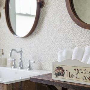 May include: A bathroom interior featuring a white sink, chrome tap, and two circular mirrors with dark wooden frames. A white box filled with rolled white towels rests on a wooden shelf. The box is labelled "Towels Hand 10¢".