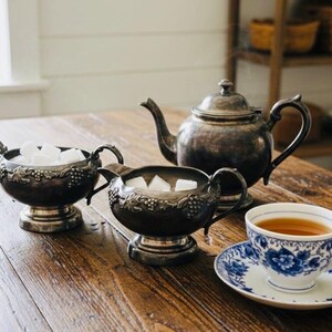 Puede incluir: Un juego de té vintage sobre una mesa de madera. El juego incluye una tetera, un azucarero, una crema y una taza de té con platillo. La taza y el platillo tienen un diseño floral azul. El azucarero y la crema están llenos de cubos de azúcar.