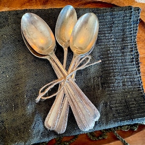 May include: A collection of vintage silver spoons, tied with twine, displayed on a dark green textured cloth. The spoons, with decorative handles, show signs of age. The spoons are a charming example of antique silverware.