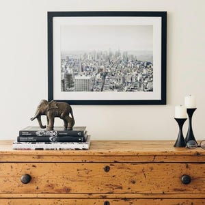 May include: A monochrome cityscape print in a black frame is displayed above a wooden chest of drawers. A detailed elephant figurine rests on books. Two black candle holders with white candles and glasses are also on display.