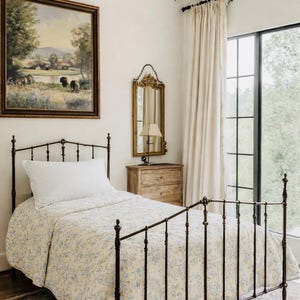 May include: A bedroom featuring a wrought iron bed with a floral quilt and white pillow. A landscape painting hangs above the bed. A gold chandelier and mirror are visible. The room has a neutral colour palette, with a window and curtains.