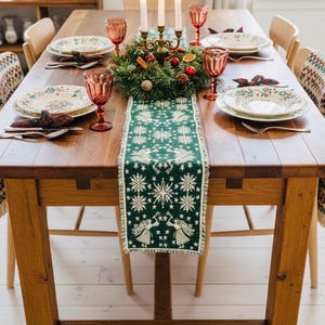 Puede incluir: Una mesa de comedor de madera preparada para una comida festiva. Un camino de mesa verde con diseños de ángeles y copos de nieve blancos decora la mesa. Platos, vasos y cubiertos están dispuestos para cada comensal, con un centro de mesa festivo.