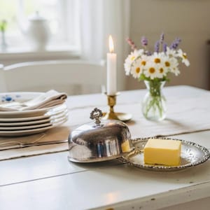 Puede incluir: Una mesa blanca con platos, una vela encendida y un pequeño jarrón con flores. Una mantequera plateada con un trozo de mantequilla sobre un plato decorativo. Una escena doméstica.