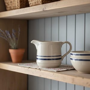 Puede incluir: Una jarra y un cuenco color crema con rayas azules están sobre una estantería de madera. Una pequeña maceta de terracota con lavanda y un paño decorativo también están en la estantería. La estantería forma parte de un mueble de madera con una pared pintada de azul claro.