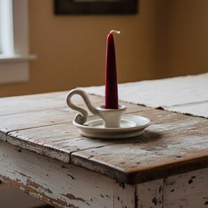 May include: A red taper candle in a white ceramic candlestick holder with a curved handle. The candle and holder sit on a distressed, white-painted wooden table. The table has a rustic, vintage appearance.