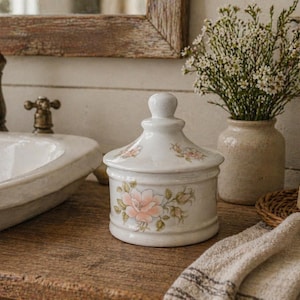 May include: A white ceramic jar with a lid, decorated with pink floral patterns. The jar is on a wooden surface, with a vase of white flowers, a soap bar, and a towel. The bathroom setting has a vintage aesthetic.