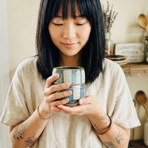 Peut inclure: Une tasse en céramique faite à la main avec un motif à rayures bleues et blanches est tenue. La tasse a une forme unique et une surface texturée. La personne porte une chemise en lin beige et a des tatouages sur les bras. L'arrière-plan comprend des étagères en bois et une décoration de cuisine.