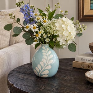 May include: A light blue ceramic vase with a white leaf pattern containing a bouquet of white daisies, blue delphinium, white hydrangea, and green eucalyptus leaves. The vase has a rounded shape and a narrow neck, holding a variety of fresh flowers.