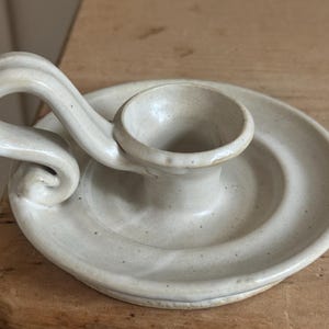 May include: Off-white ceramic candle holder with a circular base and a curved handle. The candle holder has a small cup to hold the candle and is made of speckled ceramic material.