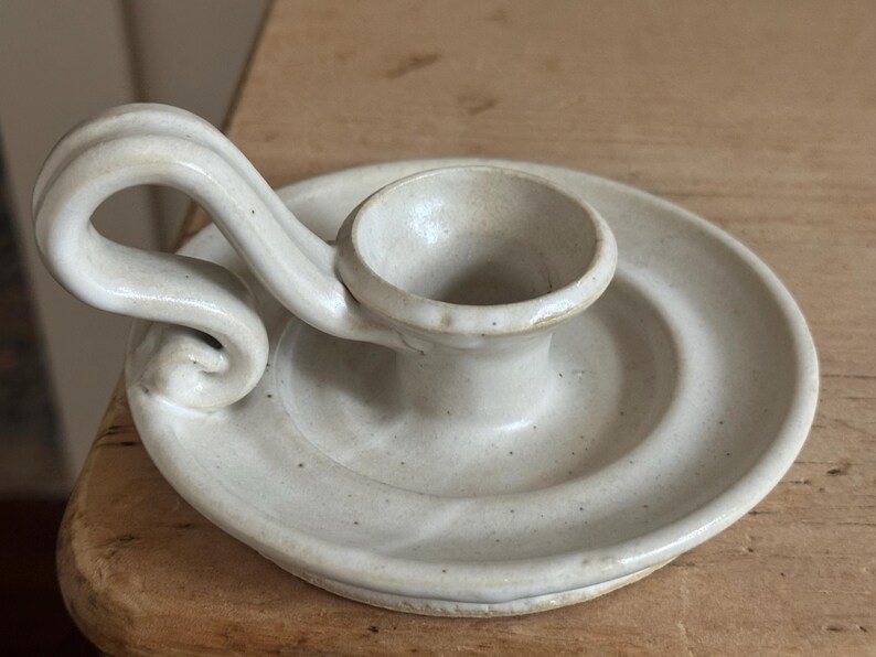 May include: Off-white ceramic candle holder with a circular base and a curved handle. The candle holder has a small cup to hold the candle and is made of speckled ceramic material.
