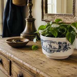 May include: A white ceramic planter featuring a blue transferware-style print of a windmill and floral motifs. The round, footed bowl contains a green pothos plant with trailing heart-shaped leaves, positioned on a rustic wooden surface.