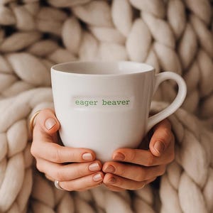 Puede incluir: Una taza de cerámica blanca con el texto "eager beaver" en letras verdes. La taza está sostenida por manos, sobre un fondo de una manta de punto grueso de color crema. La taza tiene un asa curvada.