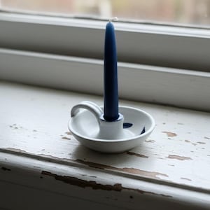 May include: A navy blue taper candle in a white ceramic candle holder with a handle. The candle holder has a small, round base with blue floral accents. The candle and holder are on a distressed white windowsill.
