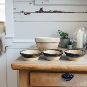 May include: A set of ceramic bowls with a cream interior and a dark brown, ribbed exterior. The bowls are displayed on a wooden surface, with a larger beige bowl behind them. Kitchenware.