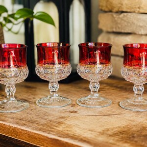 May include: Four vintage red and clear crystal wine glasses. The glasses have a red top and a clear, patterned base. They are displayed on a wooden surface.
