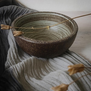 May include: A brown ceramic bowl with a speckled exterior and a light green and brown interior. The bowl sits on a grey fabric, with dried stems and seed pods resting on the bowl and fabric.