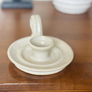May include: A ceramic candle holder with a circular base and a handle. The candle holder is a light beige color and has a small cup in the center to hold a candle. The handle curves upward.