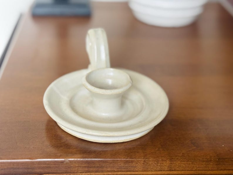 May include: A ceramic candle holder with a circular base and a handle. The candle holder is a light beige color and has a small cup in the center to hold a candle. The handle curves upward.