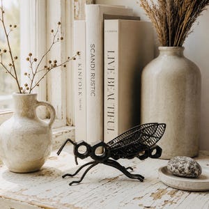 May include: A black metal insect sculpture with a mesh body and geometric head. It is displayed on a distressed white surface, alongside ceramic vases and books. The books read "SCANDI RUSTIC" and "LIVE BEAUTIFUL".