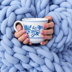 May include: A white teacup with blue floral patterns is held by hands with pink nail polish, set against a light blue chunky knit blanket. The blanket's thick, woven texture creates a cosy, warm aesthetic.