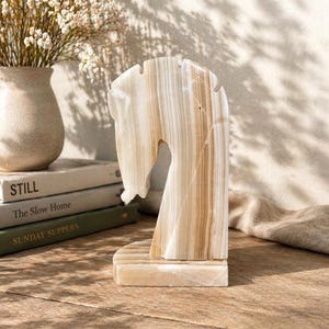 Peut inclure: Une sculpture décorative en forme de tête de cheval sculptée dans de la pierre beige et marron rayée. La sculpture est exposée sur une surface en bois, avec des livres et un vase de fleurs en arrière-plan. Un objet de décoration unique.