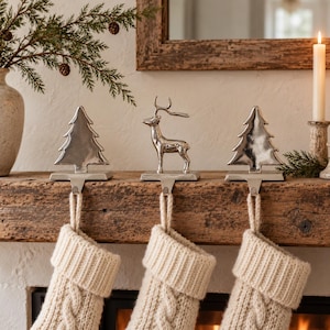 May include: Three silver-toned metal stocking holders featuring two stylised evergreen trees and one standing deer figure. Each holder has a flat base and a hook extending downward, currently supporting three cream-coloured knit stockings.