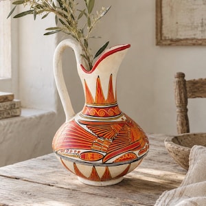May include: A ceramic pitcher with a cream-coloured base featuring hand-painted orange and red bird motifs, geometric patterns, and a curved handle. The vessel has a wide, rounded body and a flared spout, containing a green leafy branch.