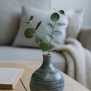 May include: A small, blue and white striped ceramic vase holds sprigs of eucalyptus with round, green leaves. The vase sits on a light wood table, with a book and a sofa in the background.