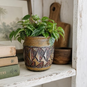 May include: A cylindrical ceramic planter with a mustard yellow glaze and a dark, textured band featuring a repeating geometric diamond pattern in relief. The planter contains a lush green plant with variegated heart-shaped leaves.