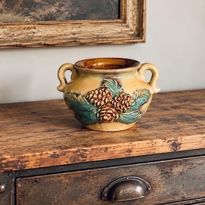 May include: A ceramic pot with two handles, featuring a tan base colour and a raised relief design of brown pinecones and green pine needles on the front. The interior of the pot is glazed in a deep brown, and the exterior has a glossy finish.