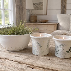 May include: A white ceramic bowl filled with green foliage sits beside two white ceramic candle holders. Each candle holder features a textured surface with green botanical illustrations and a lit candle wick centred inside the vessel.