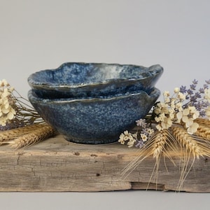 May include: Two blue speckled ceramic bowls with a wavy rim are stacked on a weathered wooden surface. Dried wheat and white and purple flowers are arranged around the bowls, creating a rustic, natural aesthetic.