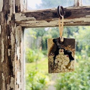 May include: A rectangular ceramic ornament with a textured, mottled surface featuring shades of cream, brown, and black. The piece has a circular hole at the top, threaded with a thin, light-coloured raffia string for hanging purposes.