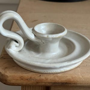 May include: Off-white ceramic candle holder with a circular base and a curved handle. The candle holder has a small cup to hold a candle and is made of a speckled, textured ceramic material. The design is simple and elegant.