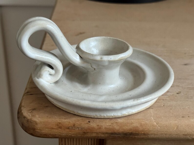 May include: Off-white ceramic candle holder with a circular base and a curved handle. The candle holder has a small cup to hold a candle and is made of a speckled, textured ceramic material. The design is simple and elegant.
