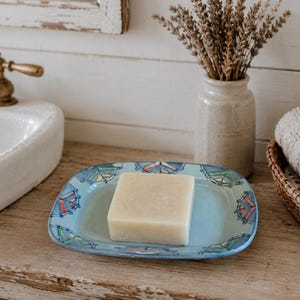 May include: A square, light blue ceramic soap dish with a hand-painted border featuring geometric patterns in blue, green, and orange. A solid, rectangular, off-white bar of soap sits centered on the dish, which has slightly raised, rounded edges.
