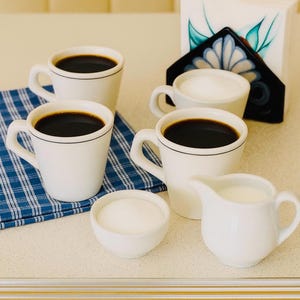 May include: A collection of white coffee cups filled with dark coffee, alongside a small creamer and a bowl of sugar. A blue and white checked napkin and a floral napkin holder are also visible.