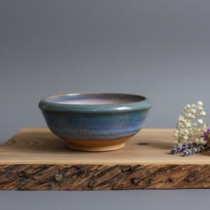 May include: A handcrafted ceramic bowl with a blue and purple glaze. The bowl is displayed on a wooden board with a natural edge. Dried flowers and a seed pod are arranged nearby. The bowl's diameter is approximately 15 cm.