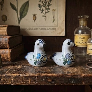 May include: Two ceramic bird ornaments with white bodies, blue and yellow floral patterns, and blue detailing. They sit on a wooden surface with old books, bottles, and a thimble in the background.