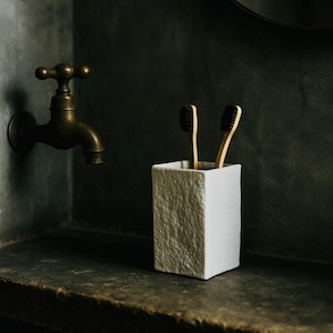 May include: A white, textured toothbrush holder containing two bamboo toothbrushes. The holder is on a dark countertop near a vintage brass tap. The bathroom has a dark, moody aesthetic.