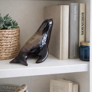 May include: A glossy, dark brown ceramic seal figurine with a smooth, reflective surface. The sculpture is positioned on a white shelf, alongside books and decorative items, creating a coastal or nautical theme.