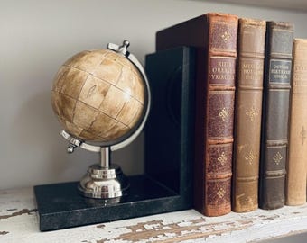 May include: A vintage-style globe with a cream-colored surface and a silver stand sits on a black marble bookend. Several antique books with leather-bound covers in shades of brown and tan are arranged on a distressed white shelf, creating a classic aesthetic.