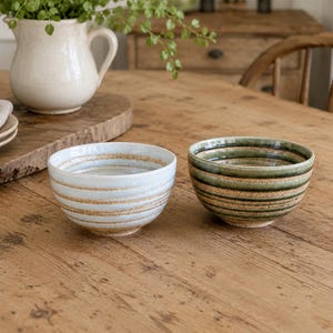 May include: Two ceramic bowls with a rounded shape and horizontal ribbed texture. One bowl features a light blue glaze with exposed tan clay, while the other has a deep green glaze with similar tan horizontal stripes around the exterior.