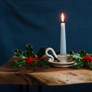 Peut inclure: Une bougie bleu pâle allumée dans un porte-bougie en céramique blanche avec une poignée décorative. La bougie est posée sur une surface en bois, ornée de houx et de baies rouges, sur fond bleu foncé. Festif et saisonnier.