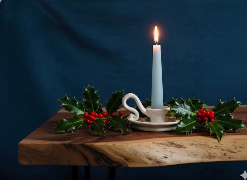 May include: A lit, pale blue taper candle in a white ceramic holder with a decorative handle. The candle sits on a wooden surface, adorned with holly and red berries, against a dark blue backdrop. Festive and seasonal.